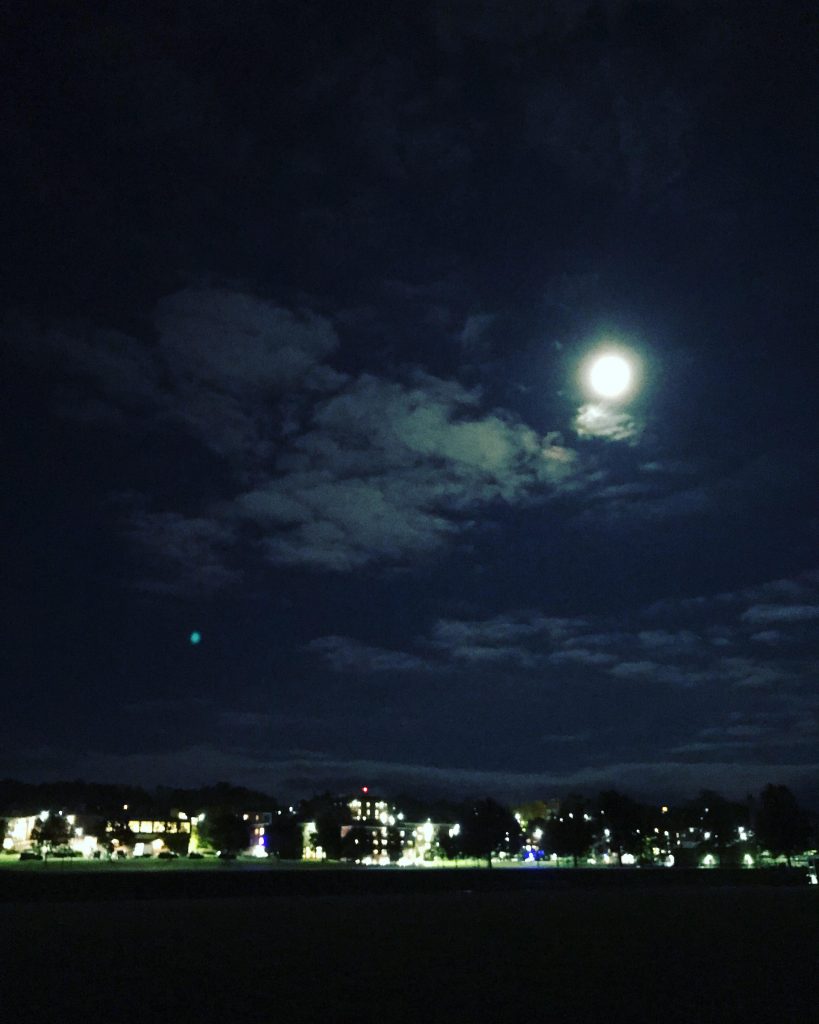 Under dark skies, a full moon appears between clouds with the SUNY Geneseo campus visible in background 