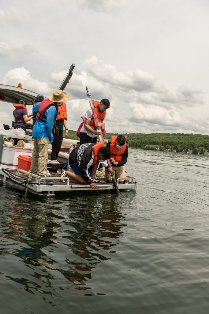 students on the edge of the boat wearing life vests pulling in a collected core from the lake